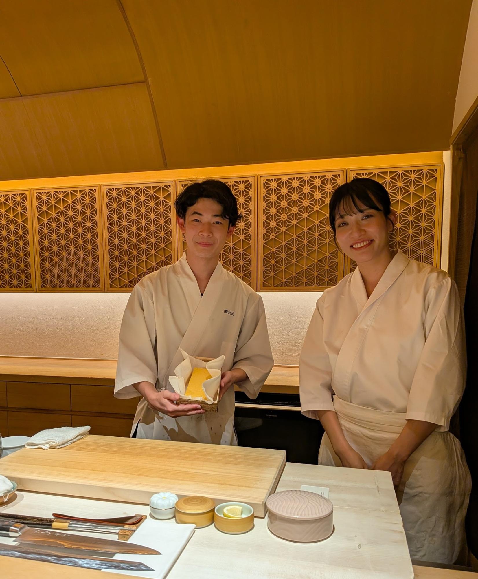 白木のカウンター越しに、名物の玉子焼きを手にして柔らかい笑顔を向ける鮨職人の井上ゆり氏と同期の男性職人